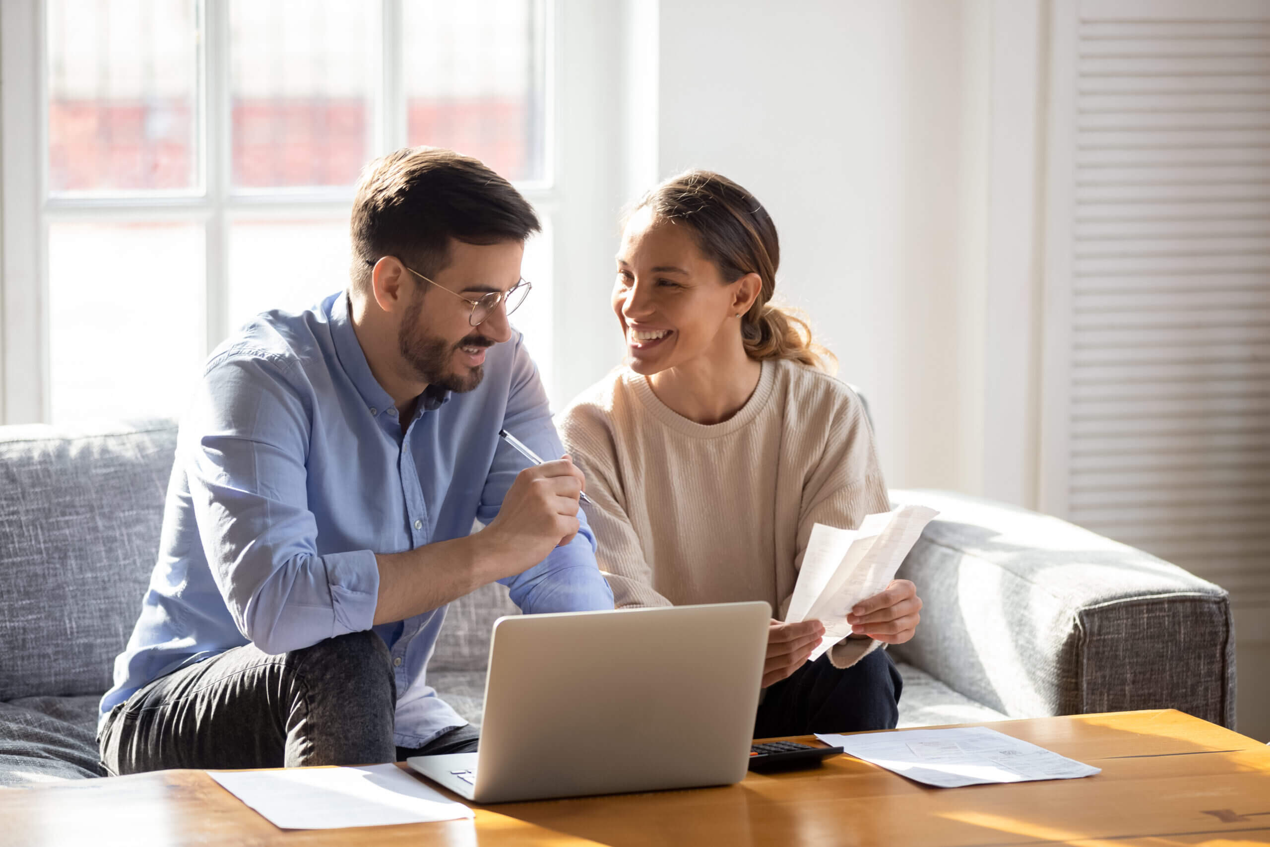 A man and woman sit on a sofa, using a laptop and examining paperwork over a coffee table.